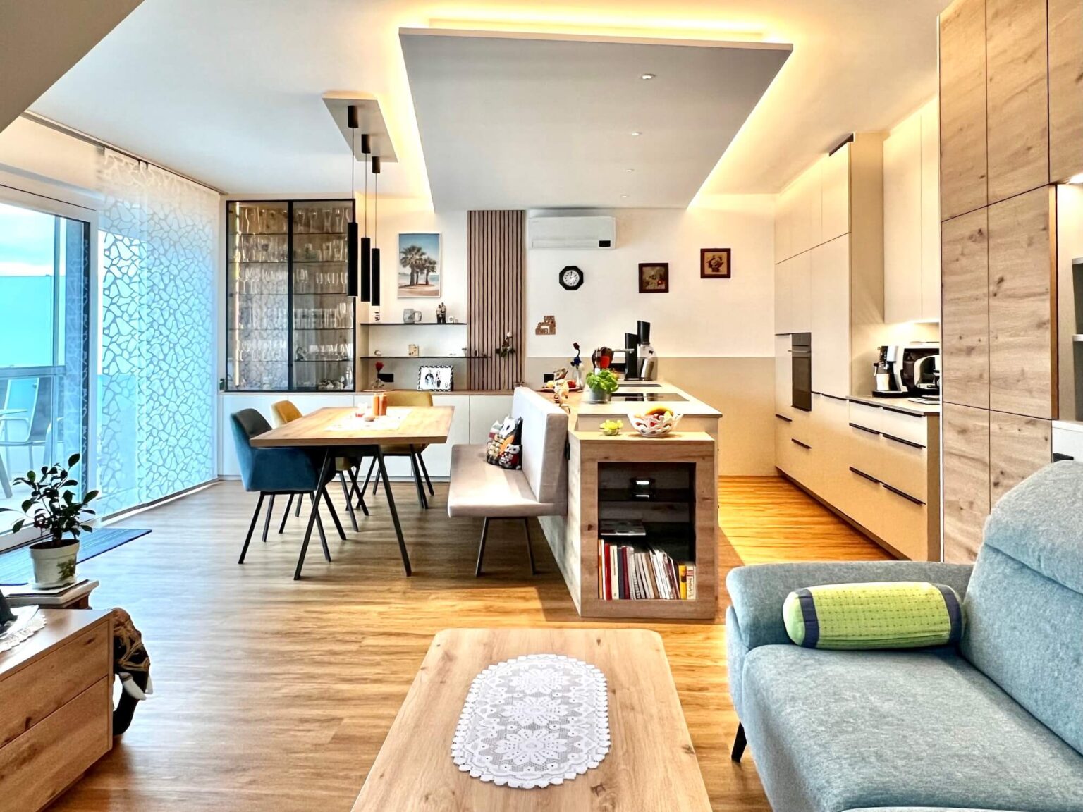 Offener Wohnraum mit einer Küchenzeile aus betongrauem und hellem Holz, Essbereich mit Tisch und Stühlen in neutralen Farben.