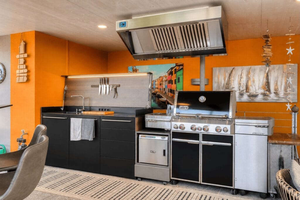 Küche im Freien mit schwarzen Küchenschränken, Edelstahl-Grill und orangefarbener Wandgestaltung, ergänzt durch Holz-Dekoration.