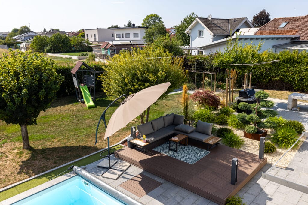 Garten mit Lounge-Bereich, Schwimmbecken, Rasenfläche, Holzterrasse, Spielplatz und bepflanzten Beeten.