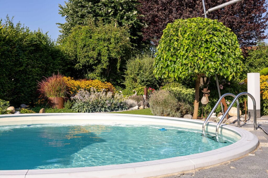 Runder Pool mit Edelstahlleiter, umgeben von vielfältiger Bepflanzung, darunter bunte Blumen und Sträucher in einem Gartenbereich.