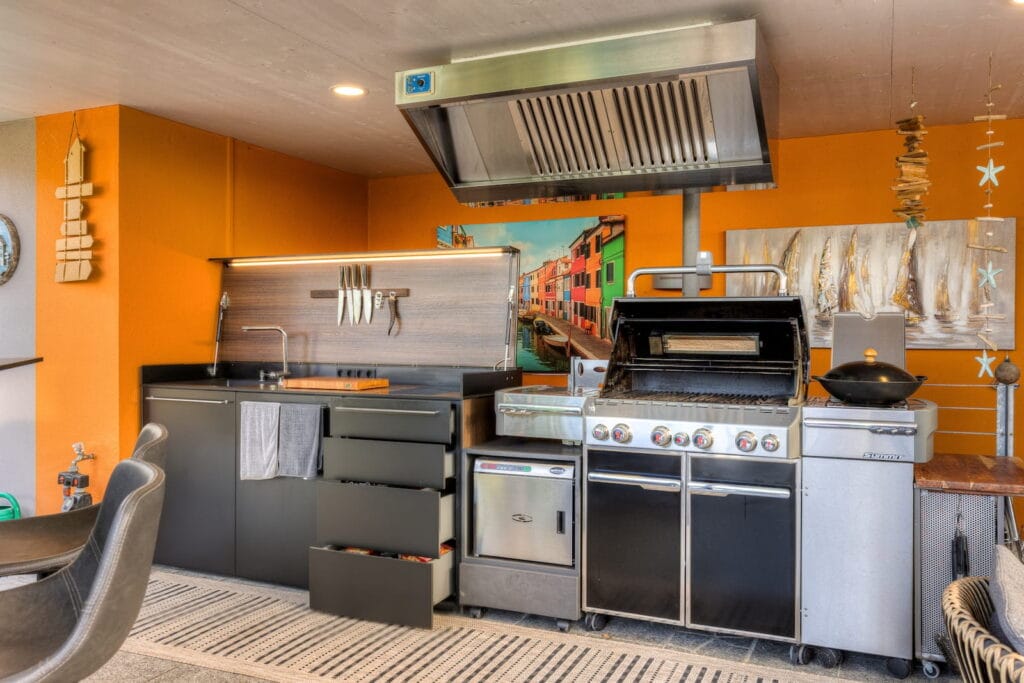 Außenküche mit schwarzer Küchenzeile, Edelstahl-Grill und orangefarbener Wand, ergänzt durch dekorative Elemente und Teppich.
