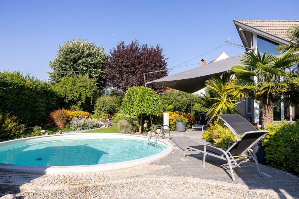 Swimmingpool im Garten, umgeben von bepflanzten Beeten und Sonnenliegen auf einer Terrasse aus Steinplatten.