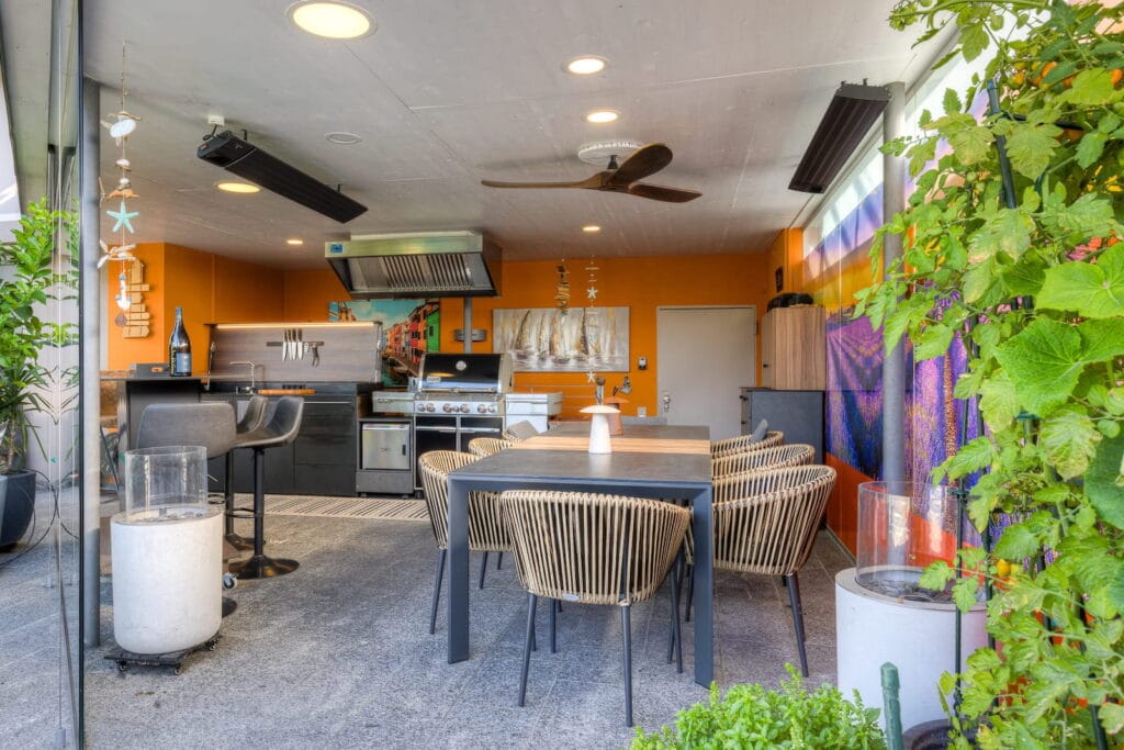 Der Außenbereich umfasst eine moderne Küche mit Grill, einem Esstisch und Stühlen aus Rattan in einer orangefarbenen Wandgestaltung.