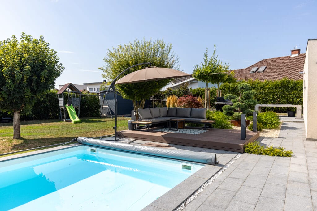Garten mit Pool, Holzterrasse, Lounge-Möbeln und Spielgerät, umgeben von Bäumen und Büschen in Marchtrenk.