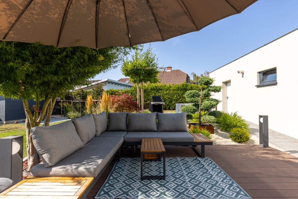 Gemütliche Terrasse mit grauem Sofa, Holztablett, Teppich und bepflanzten Beeten unter einem großen Sonnenschirm.