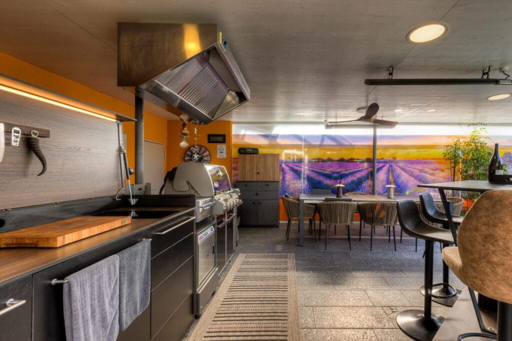 Küche und Lounge-Bereich mit Holzbelegen, Metallgrill, runder Esstischgruppe und einer Wand mit Lavendelmotiv.
