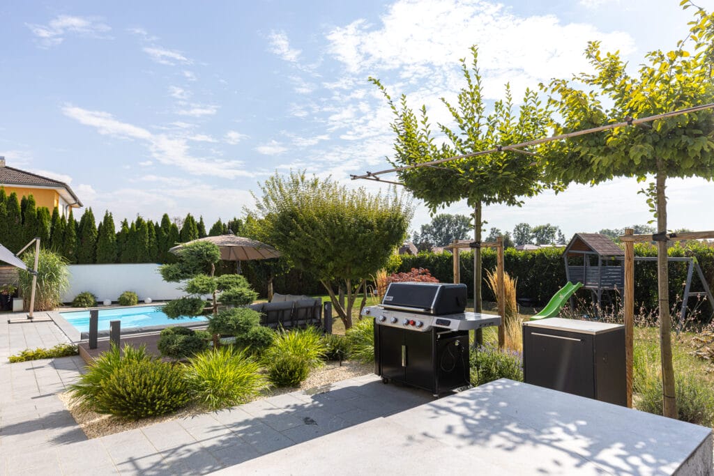 Der Garten mit Pool, Grill, Sitzgelegenheiten und Bäumen ist umgeben von schlichten, hellen Steinplatten und begrünten Beeten.