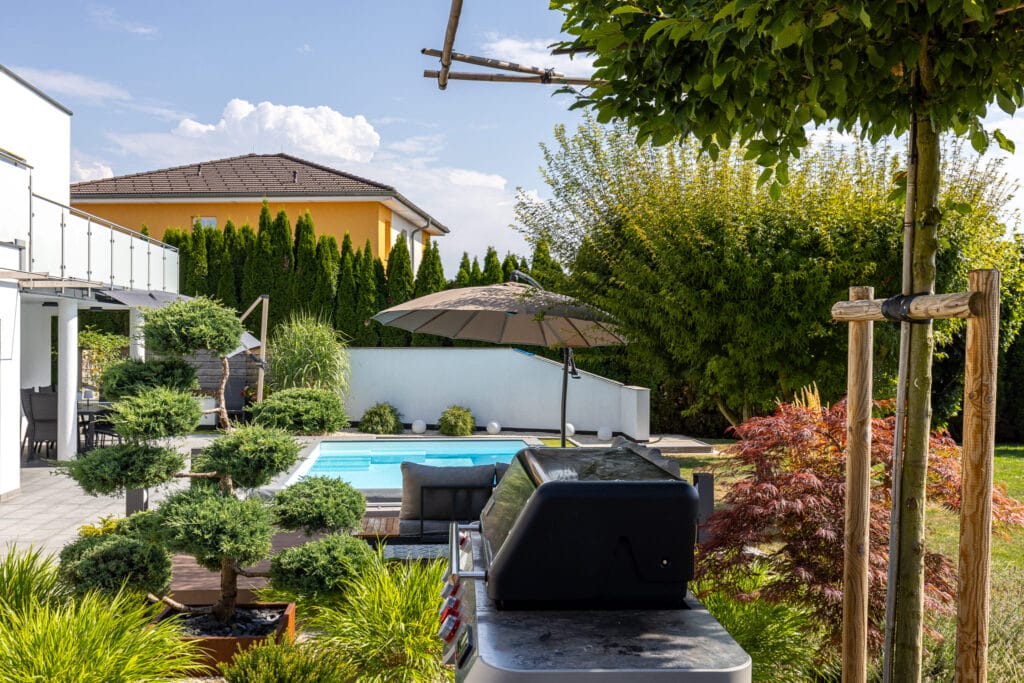 Freifläche mit Swimmingpool, Grill, Sonnenschirm und gepflegten Pflanzen, umgeben von hohen Hecken und Bäumen.