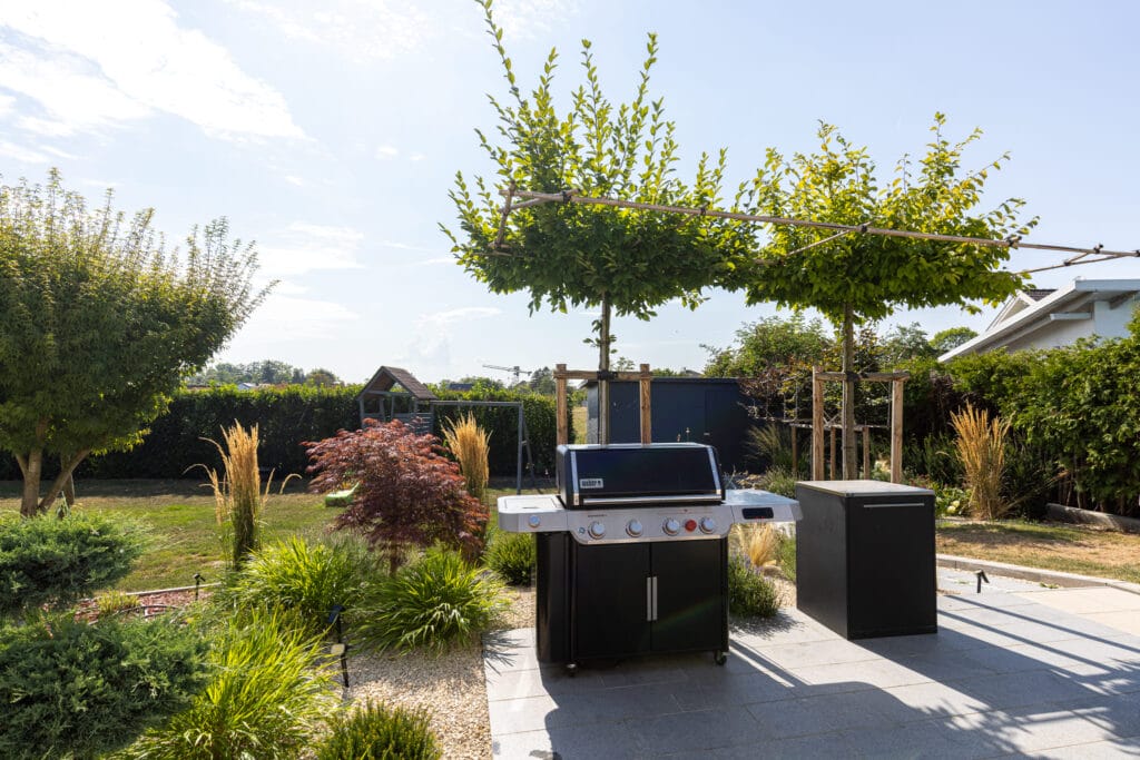 Terrasse mit Grill, umgeben von Bäumen und Gartenpflanzen, gepflastert mit grauen Steinen.