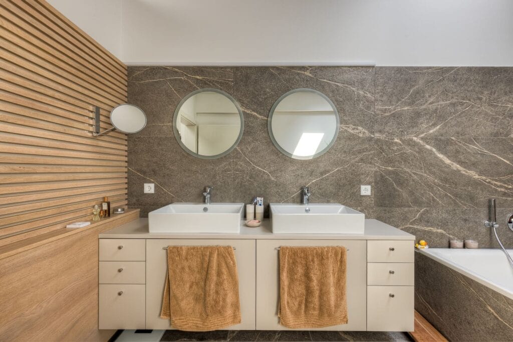 Modernes Badezimmer in einer hellen Dachgeschosswohnung mit grauen Fließen, beigen Badezimmermöbeln und runden Spiegeln.