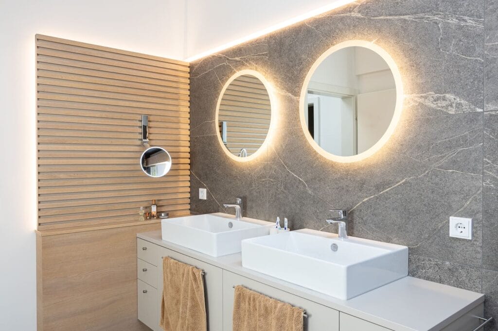 Zweibad mit Doppelwaschbecken, runden Spiegeln und einer Wand aus grauem Stein, ergänzt durch Holzverkleidungen und beiges Mobiliar.