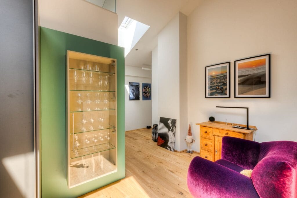 Dachgeschosswohnung mit Holzfußboden, grüner Wandnische für Gläser, violettem Sessel und Bilderrahmen an der Wand.