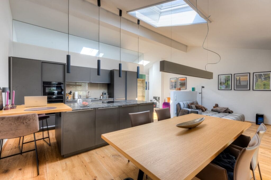 Küche und Wohnbereich in einer Dachgeschosswohnung mit hellen Wänden, Holzfußboden, modernen Möbeln und einer großen Fensterfläche.