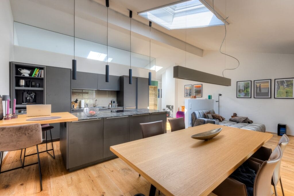 Küche und Wohnbereich in einer Dachgeschosswohnung mit hellen Wänden, Holzfußboden, modernen Möbeln und einer großen Fensterfläche.