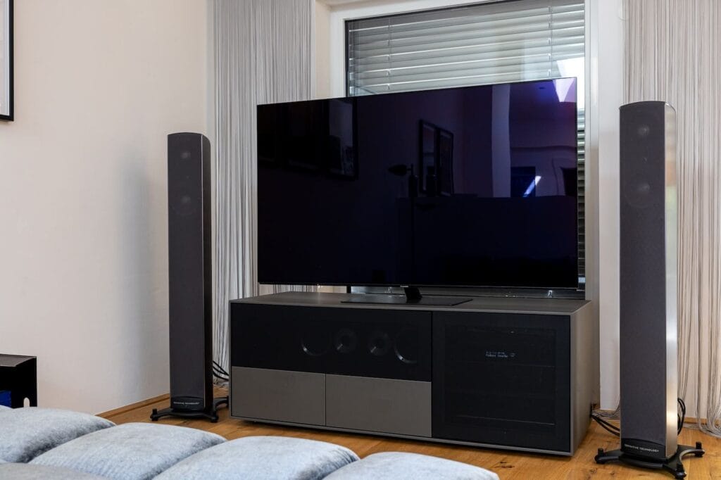 Wohnzimmer mit Holzboden, einem großen Fernseher auf einem schwarzen TV-Möbel und zwei Lautsprechern neben dem Fenster.
