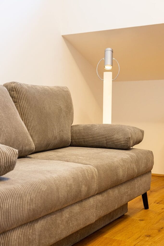 Ein Raum mit einem beigen, gestreiften Sofa und einer modernen Stehlampe vor einer hellen Wand und einem Holzboden.