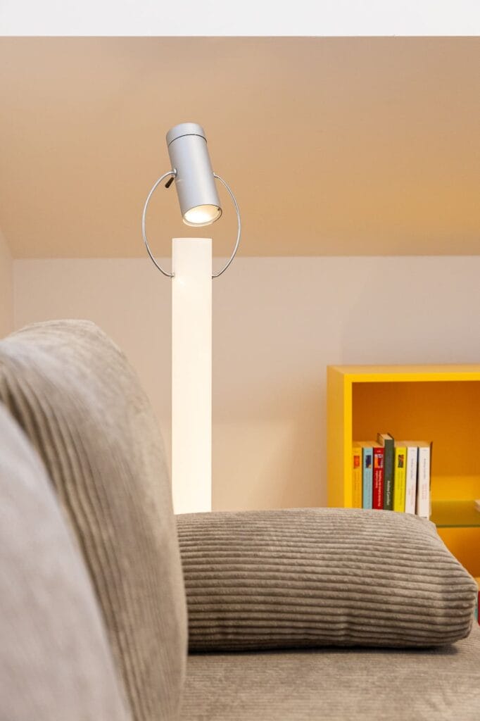 Ein Raum mit einem beigen, gestreiften Sofa und einer modernen Stehlampe vor einer hellen Wand. Im Raum steht ein gelbes Bücherregal.