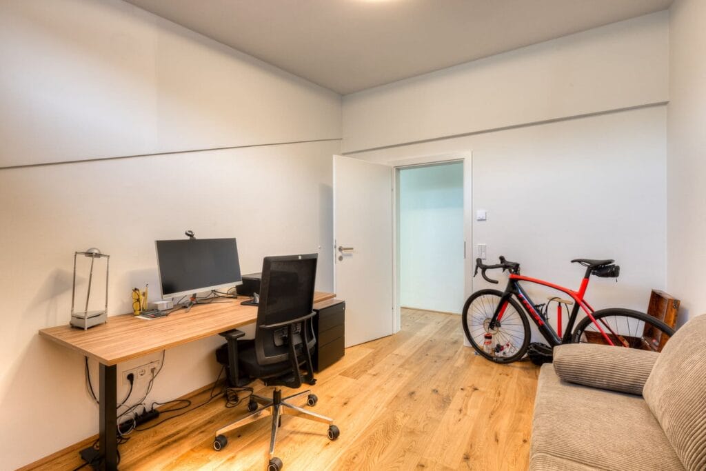 Büro in einer Dachgeschosswohnung, helle Wände, Holzfußboden, Schreibtisch mit Monitor und schwarz-rotem Fahrrad in der Ecke.