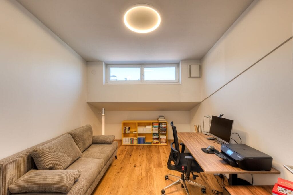 Büro in einer Dachgeschosswohnung, helle Wände, Holzfußboden, Schreibtisch mit Monitor, einer beigen Couch und einem gelben Bücherregal.