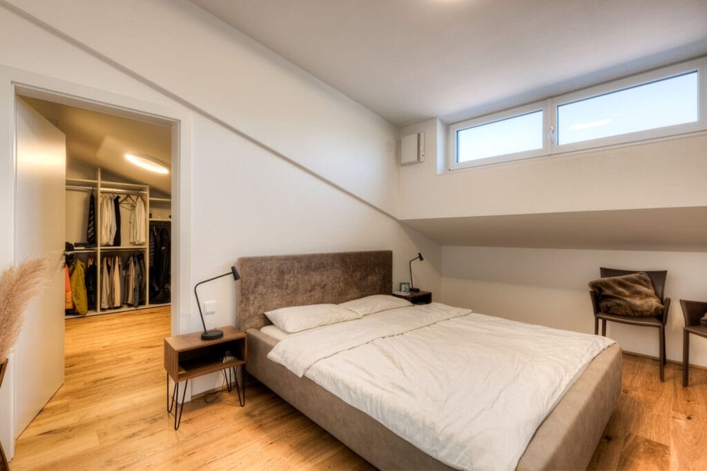 Schlafzimmer in einer Dachgeschosswohnung mit Holzfußboden, einem Bett mit beigefarbenem Kopfteil und einer kleinen Nachttischlampe. Eine Tür führt ins Ankleidezimmer.