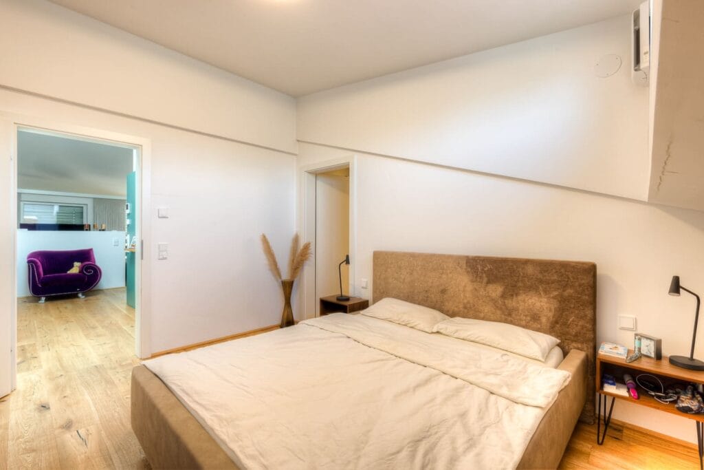 Schlafzimmer in einer Dachgeschosswohnung mit Holzfußboden, einem Bett mit beigefarbenem Kopfteil und einer kleinen Nachttischlampe.