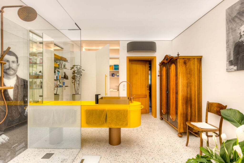 Badezimmer mit gelber Waschtischanlage, Glasdusche, terrazzo Boden und Holzschränken, ergänzt durch dekorative Pflanzen.