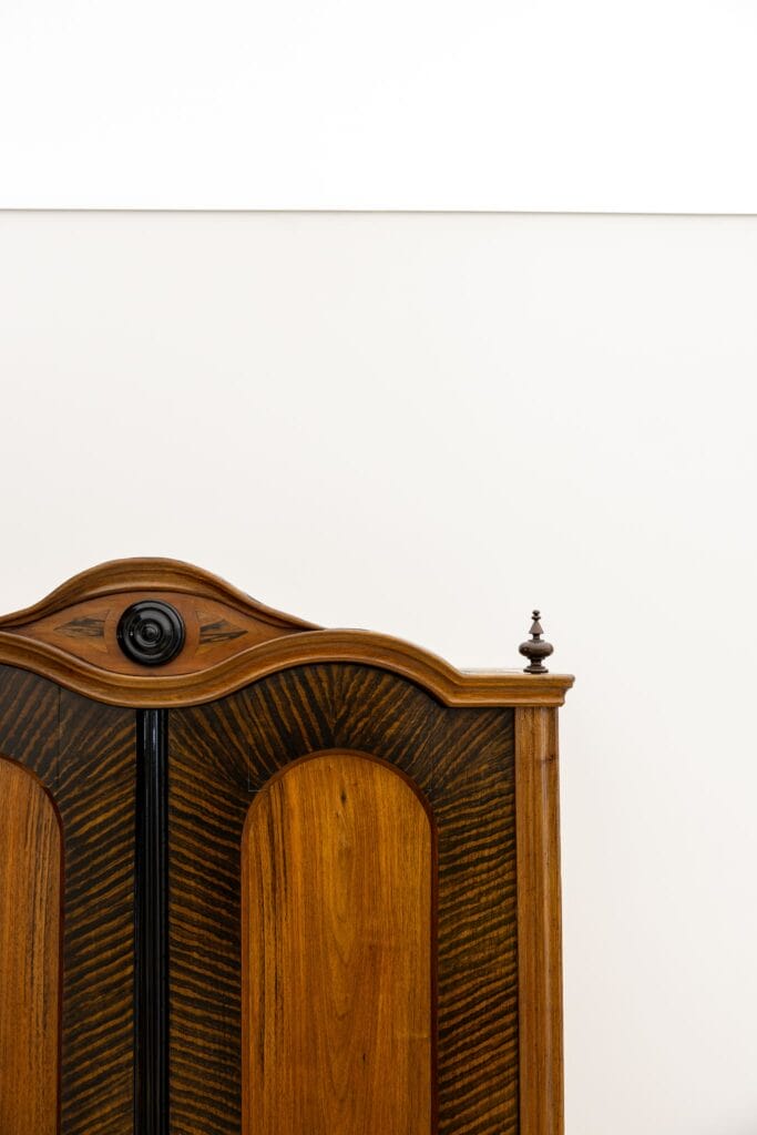 Ein hölzerner Schrank mit geschwungenem Oberteil, hellen und dunklen Holzflächen, vor einer weißen Wand.