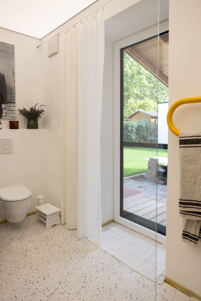 Badezimmer mit hellem Terrazzo-Boden, weißen Wänden, großer Fenstertür und gelben Handtuchhalter.
