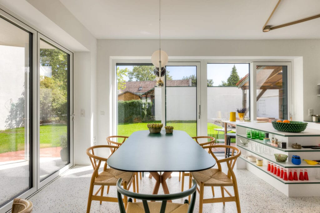 Esszimmer mit ovalem Tisch und Holzstühlen, große Fenster zur Gartenansicht, helle Wände und moderne Regale.