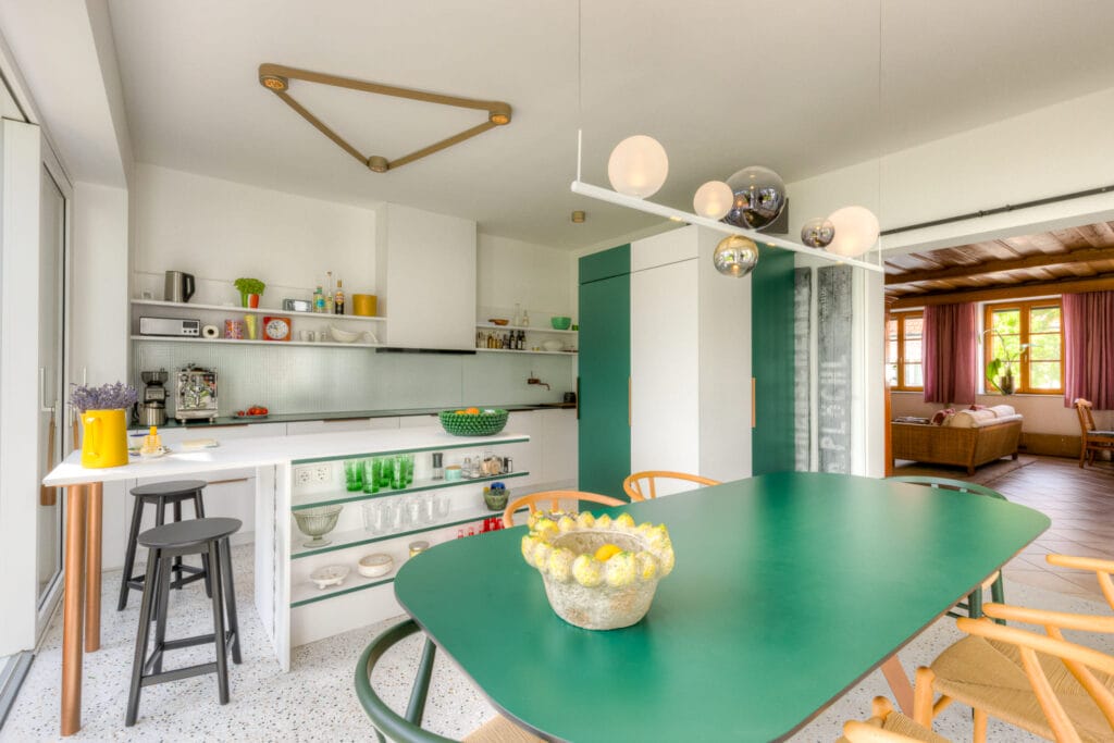 Moderne Küche mit grüner Esstischfläche, weißen Möbeln, hellem Boden und offenen Regalen, angrenzendem Wohnzimmer mit Holzdetails.