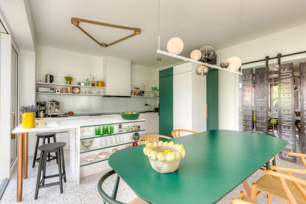 Küche mit grünem Tisch, offenen Regalen, modernen Stühlen und Teppichboden, sowie einem großen Fenster und einer Lichtinstallation.