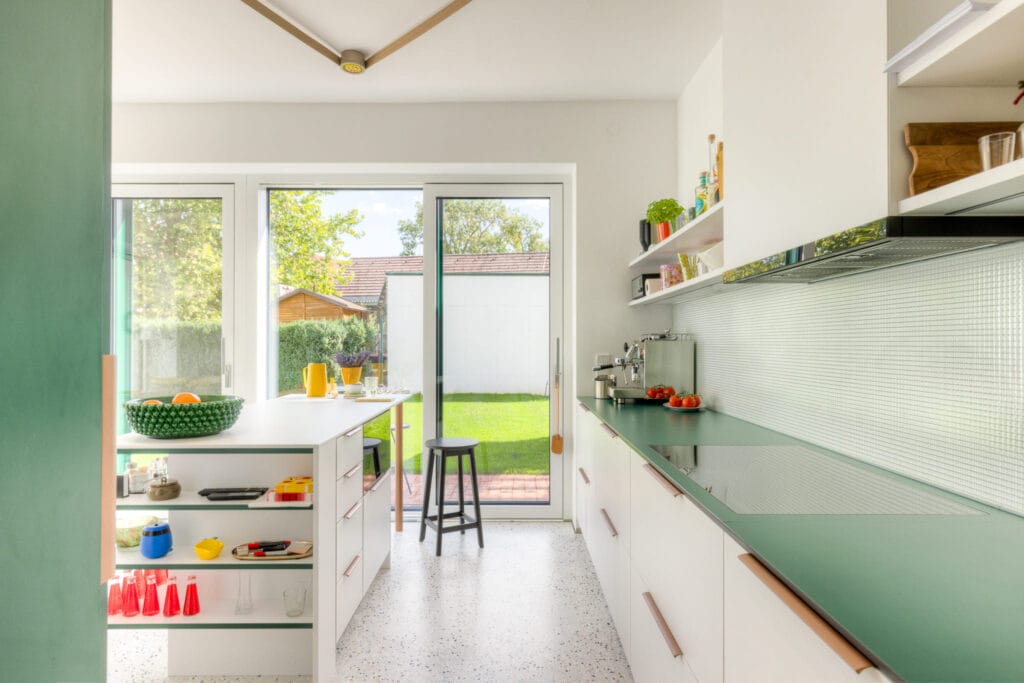 Moderne Küche mit grüner Arbeitsfläche, weißen Möbeln, offenen Regalen und großen Fenstern zum Garten.