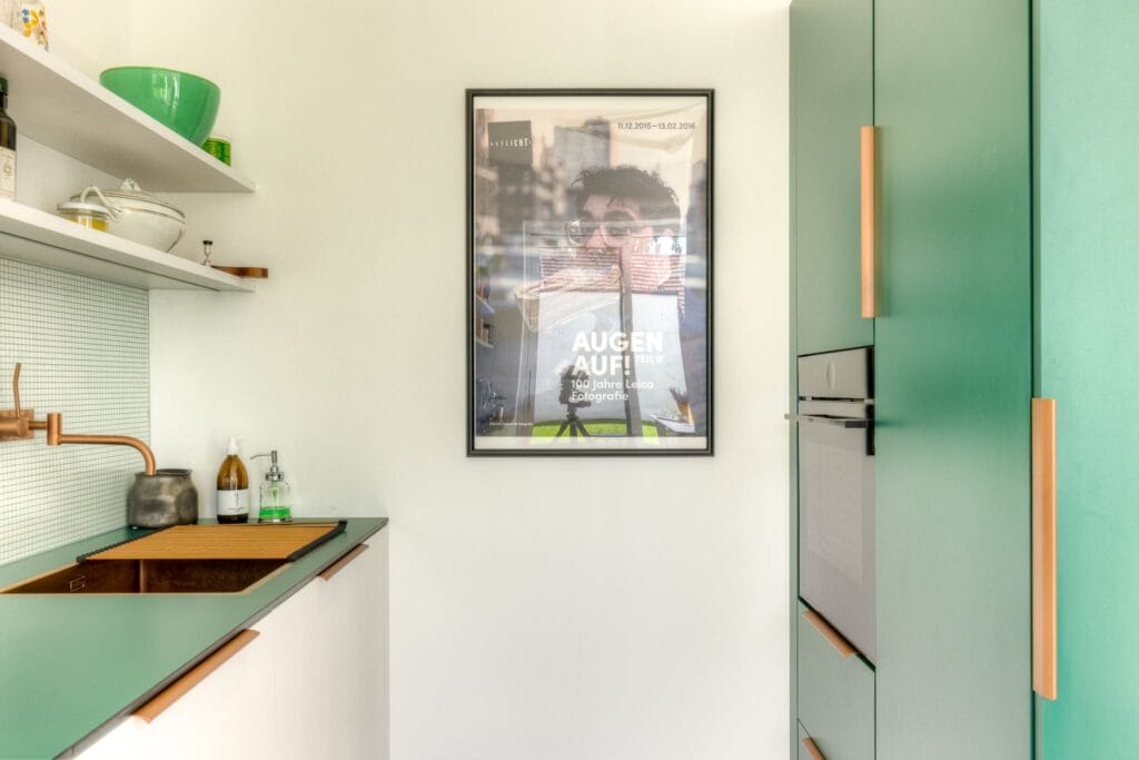 Küche mit grünen Oberflächen, weißen Schränken, Kupferarmaturen und einem gerahmten Kunstwerk an der Wand.