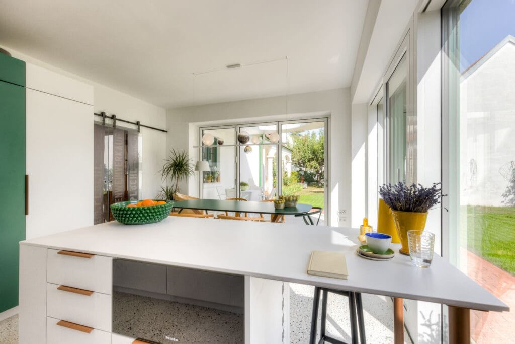 Helle Küche mit weitem Fenster, grünen Akzenten, weißem Arbeitsbereich, Esstisch und Blick auf den Garten.
