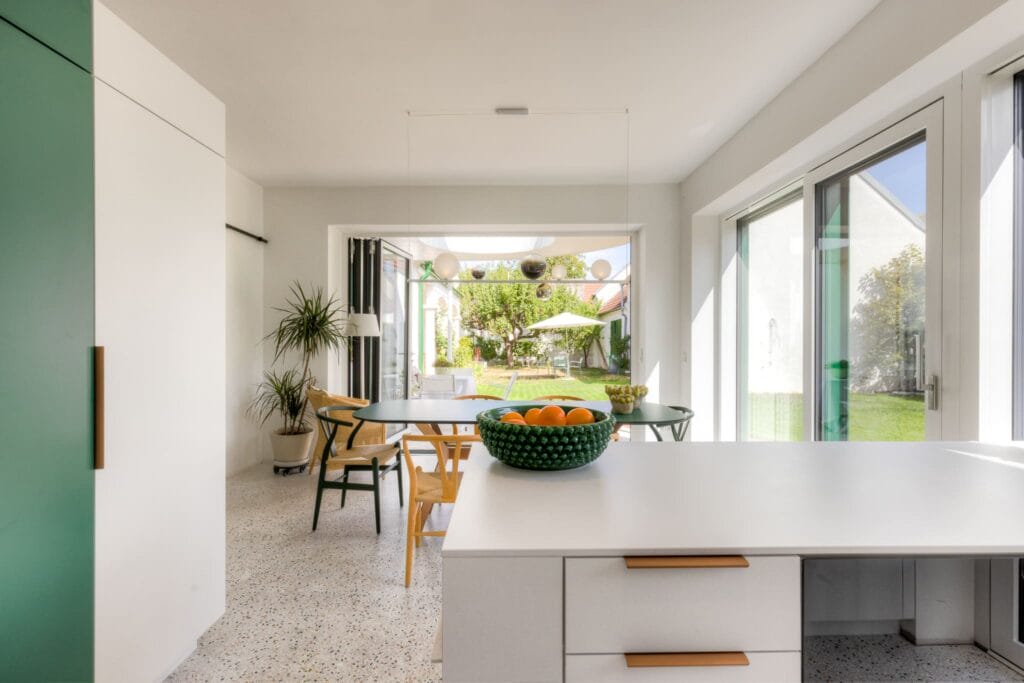 Helle Küche mit weißem Küchenblock, grünen Akzenten, Terrassenblick und Pflanzen, ergänzt durch eine Essgruppe aus Holz.