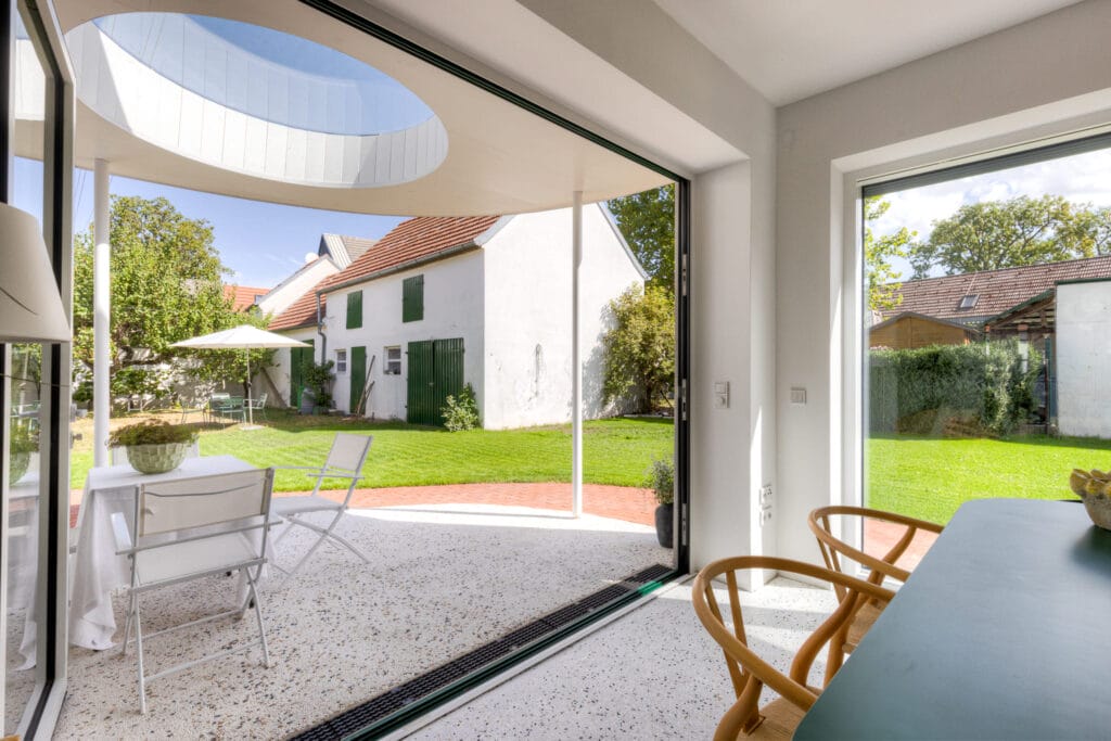 Heller Wohnraum mit großen Fenstern, weißen Wänden und einem Tisch, der auf einen Garten mit Rasen und Sonnenschirm blickt.