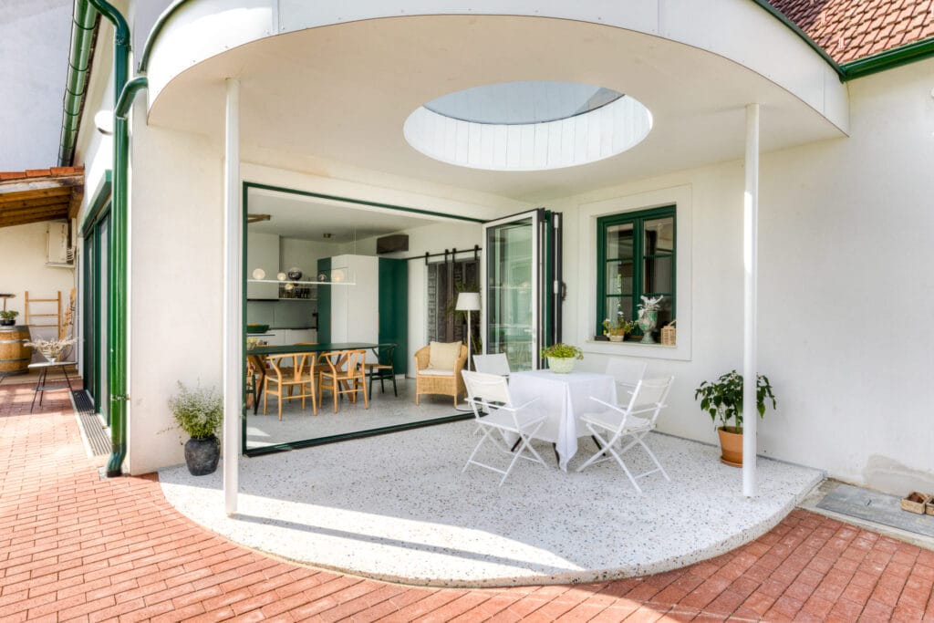 Eingang über die Terrasse zu einem hellen Wohnraum mit Glaswand, Terrazzo-Lösung für den Boden, der sich von innen nach außen zieht, grünen Fensterrahmen und einem kleinen Esstisch auf einer Terrasse.