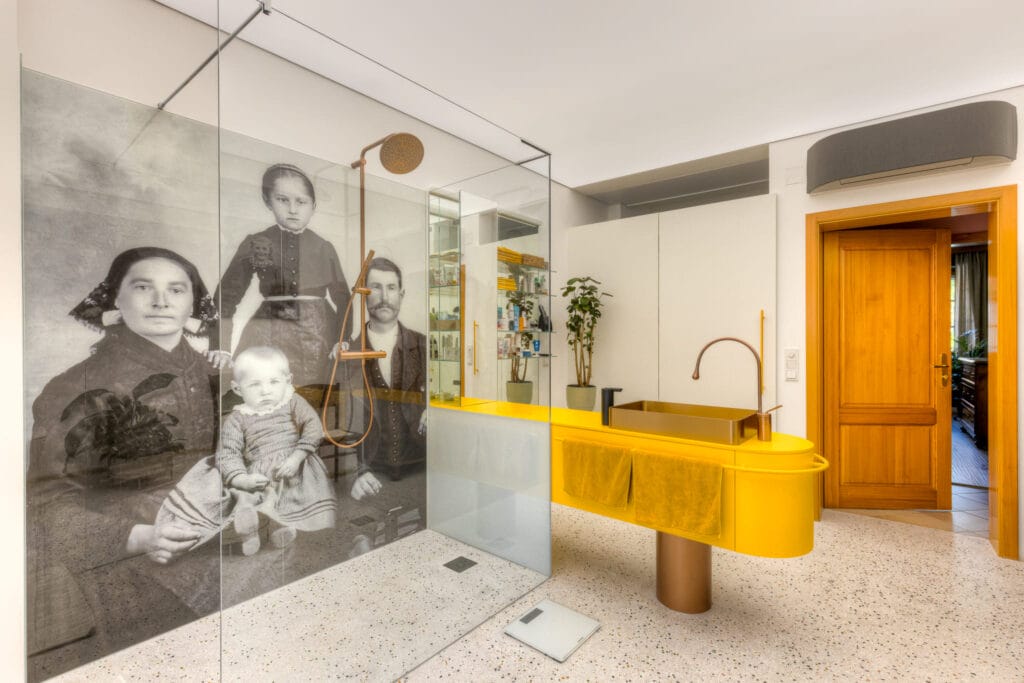 Badezimmer mit Glasduschwand, gelbem Waschtisch, terrazzo-beschichtetem Boden und vintage Familienporträts als Dekoration.