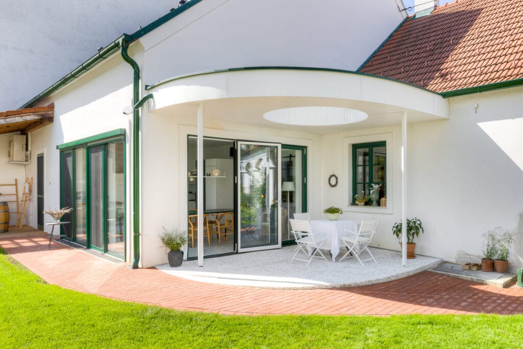 Terrasse mit runder Überdachung, verglasten Wänden, weißen Tisch und Stühlen, umgeben von Rasen und Ziegelpflaster.
