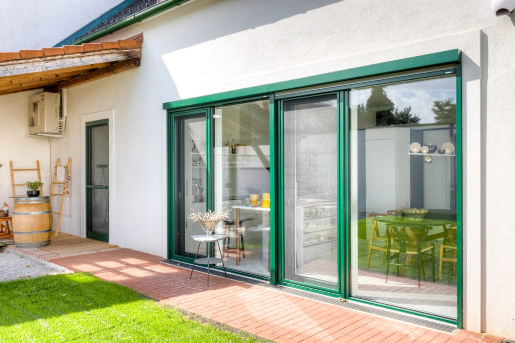 Ein hinterer Außenbereich mit großen Glasfenstern, Ziegelsteinboden, Rasenfläche und einer Holzkonstruktion über einem Holzdeck.