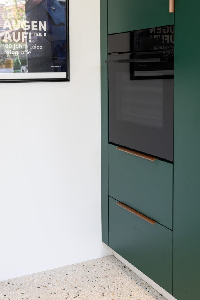 Moderne Küche mit grünen Schränken, schwarzen Geräten und einem farbigen Terrazzo-Boden. Kunstwerk an der Wand.