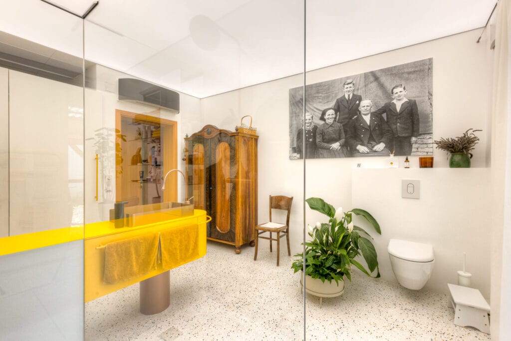 Bad mit bodengleicher Dusche, gelbem Waschbecken, Pflanzen, historischem Schrank und Schwarz-Weiß-Foto an der Wand.
