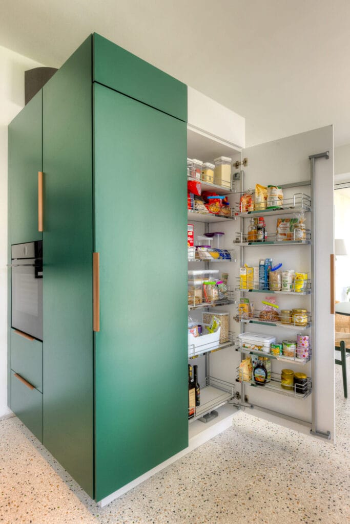 Moderne Küche mit grünem Hochglanzschrank, integriertem Backofen und offenem Regal für Vorräte auf einem terrazzoähnlichen Boden.