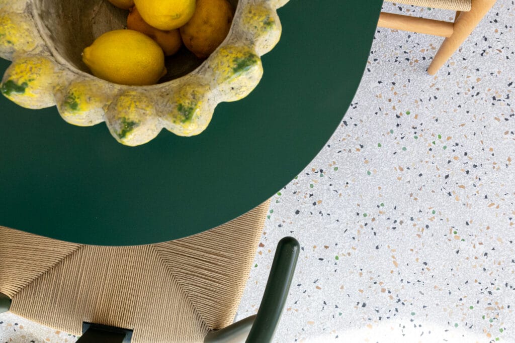 Runder Tisch mit grünem Deckel und einer keramischen Schale mit Zitronen, umgeben von einem geflochtenen Stuhl auf Terrazzo-Boden.