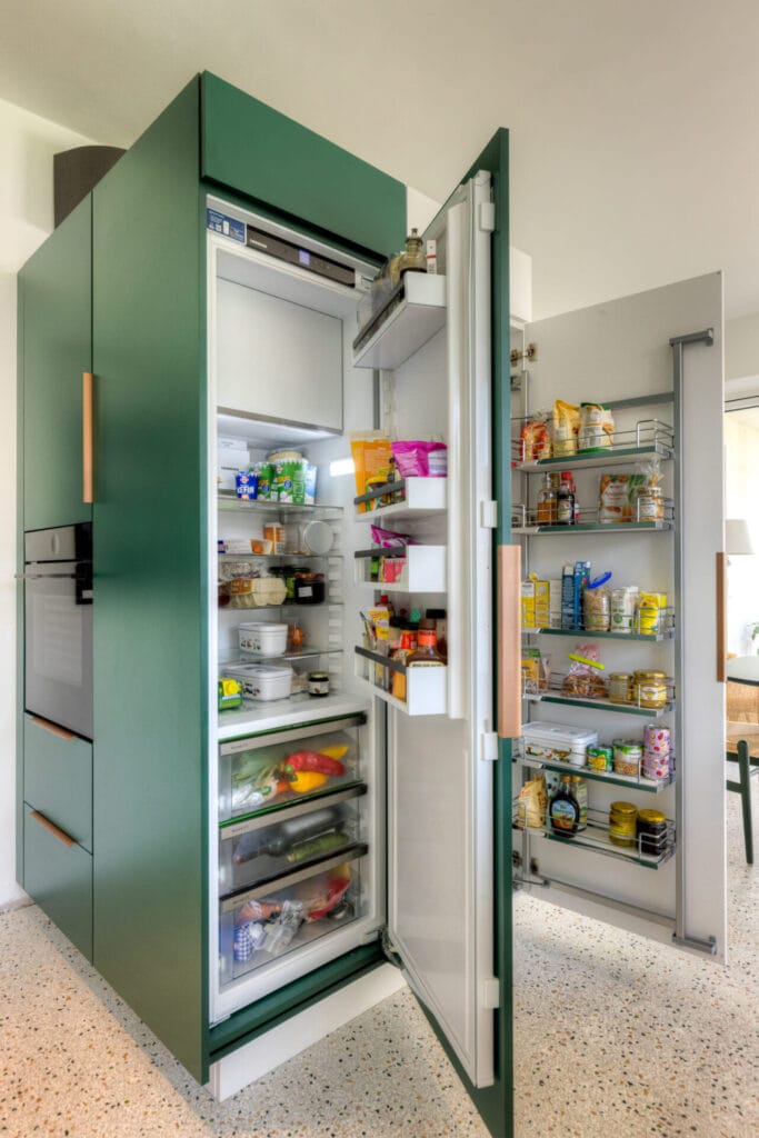 Moderne Küche mit grün-weißen Küchenschränken und Holzgriffen, Terrazzo-Boden und sitzender Pflanze neben einem Stuhl.