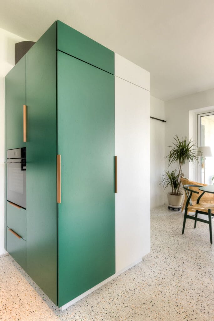 Moderne Küche mit grün-weißen Küchenschränken und Holzgriffen, Terrazzo-Boden und sitzender Pflanze neben einem Stuhl.