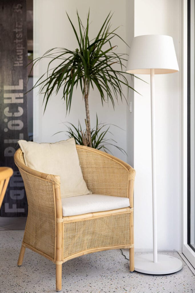 Gemütliche Sitzecke mit einem Stuhl aus Rattan, einem beigen Kissen und einer stehlampe, umgeben von einer Zimmerpflanze.