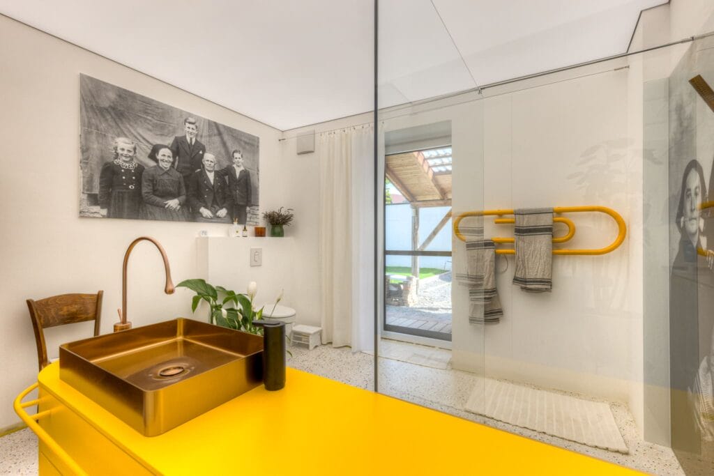 Modernes Badezimmer mit gelber Waschtischplatte, Kupferarmatur, Glasduschwand und Pflanzen, ergänzt durch ein historisches Foto.