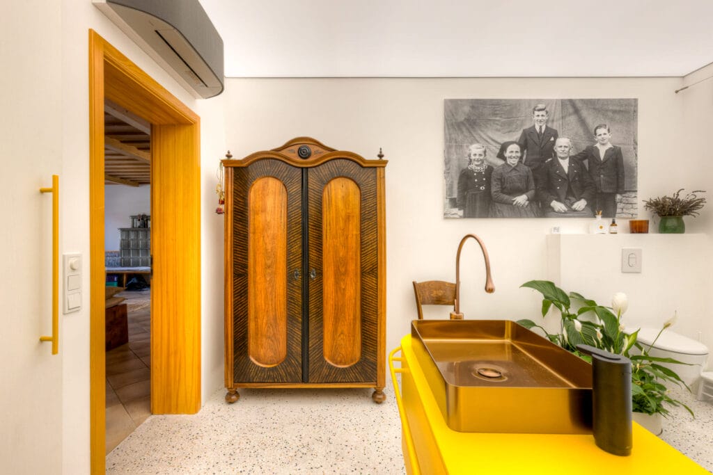 Modern trifft alt. Ein modernes Badezimmer mit gelbem Waschtisch und bronzenem Waschbecken. Dahinter ein alter, historischer Schrank.