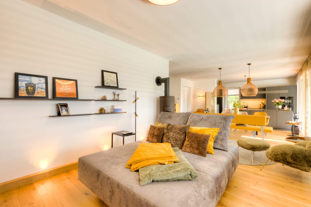 Geräumiger Wohnraum mit einem grauen Sofa, gelben Kissen, Holzfußboden und offener Küche im Hintergrund.